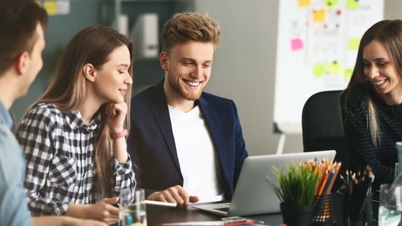 4 junge IT-Spezialisten, die in einem modernen Büro zusammen arbeiten. Sie sitzen zu viert am Tisch und blicken gemeinsam auf einen Laptop.