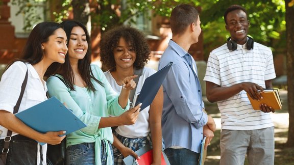 Fröhliche Studentinnen überprüfen ihren Stundenplan im Freien, während im Hintergrund Jungs miteinander sprechen – Teilnehmerinnen des Resilienz-Coachings GIP für beruflichen Erfolg bei der Organisation ihres Trainingsplans.
