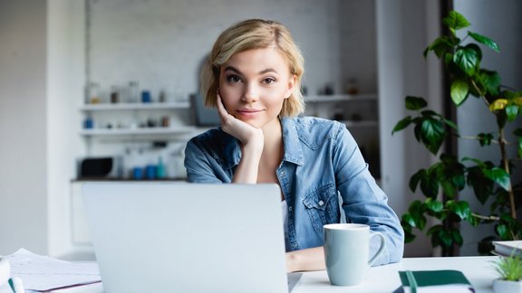 Junge Frau guckt in die Kamera und stützt dabei ihren Kopf auf ihrer Hand ab. Sie arbeitet im Homeoffice und sitzt vor einem Laptop. Daneben steht eine Kaffetasse und liegen 2 Notizbücher.