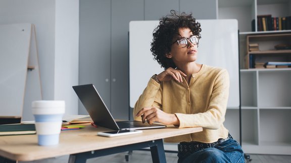 Millennial Geschäftsfrau arbeitet nachdenklich an einem Schreibtisch mit geöffnetem Laptop arbeitet. Sie trägt Brille und sitzt in einem Open-Space-Büro.