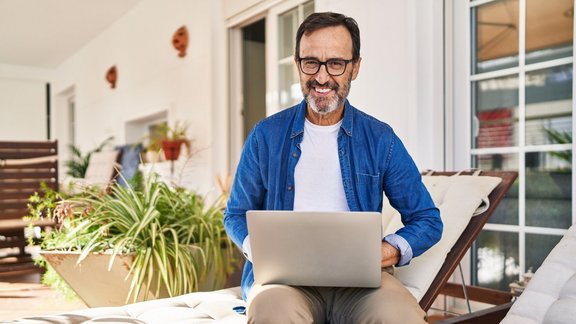 Mann mittleren Alters lächelt selbstbewusst, während er einen Laptop auf der Terrasse zu Hause benutzt. Er ist Teilnehmer des Kurses 'Wiedererwerb der Grundkompetenzen', um digitale und berufspraktische Fähigkeiten zu stärken.