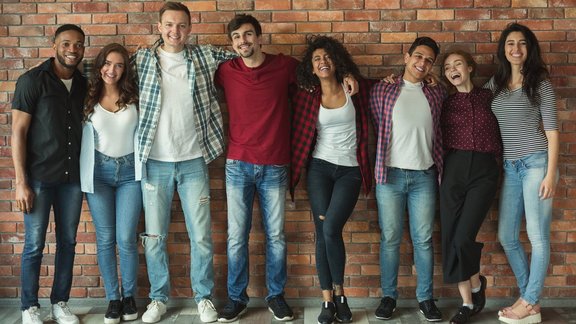 Fröhliche multikulturelle Freunde, die lachen und sich umarmen, während sie für ein Gruppenporträt posieren. Sie stehen versammelt vor einer roten Backsteinmauer.
