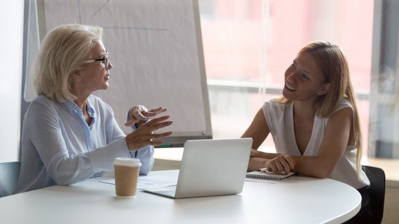 Ältere Geschäftsfrau und Mentorin spricht mit einer weiblichen Coaching Teilnehmerin im Konferenzraum, erklärt Aufgaben, diskutiert Geschäftsstrategien, Konzepte zur Mitarbeiterschulung, Brainstorming und die Beratung von Mitarbeitern und Kunden.