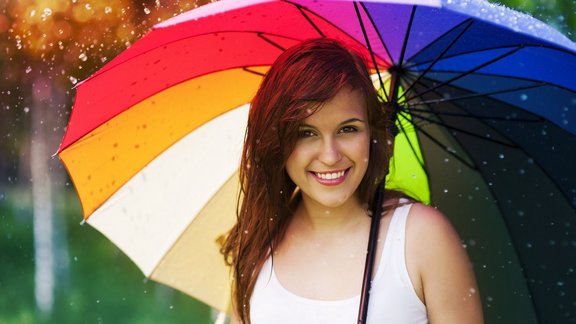 Frau steht in strömenden Regen draußen im weißen Tshirt unter einem Regenbogenfarbigen Schirm und lächelt dabei