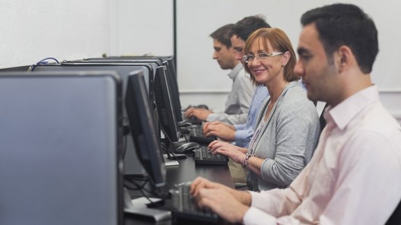 Glückliche erwachsene Studentin sitzt im Computerkurs mit anderen Teilnehmenden und lächelt in die Kamera.