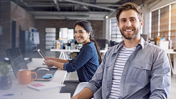Nahaufnahme eines Mitarbeiters im Büro. Hinter ihm sitzt eine Frau, beide blicken freudig in die Kamera.