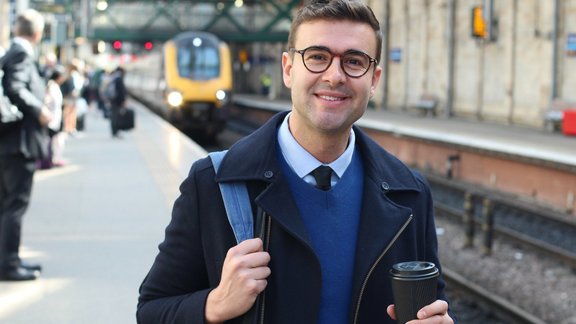 Ein Mann, der einen frühen Morgenzug zur Arbeit nehmen möchte und freudig in die Kamera lächelt. Hinter ihm sieht man den Bahnsteig an dem der Zeug einfährt.