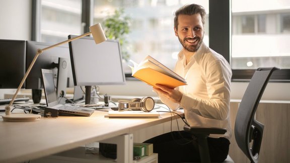 Mann im Büro sitzt vor einer Fensterfront an einem Schreibtisch mit Computer. Er hat sich umgedreht, ein Buch in der Hand und lächelt.